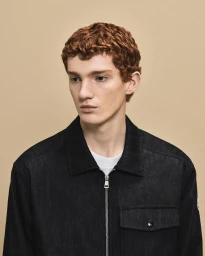 Portrait of a male model with reddish-brown curly hair wearing a black zip-up jacket against a neutral backdrop.