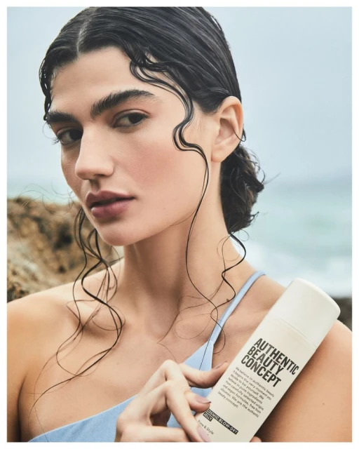 Close-up of a woman with wet hair holding a hair product on a rocky beach with ocean in the background, promoting beauty and haircare.
