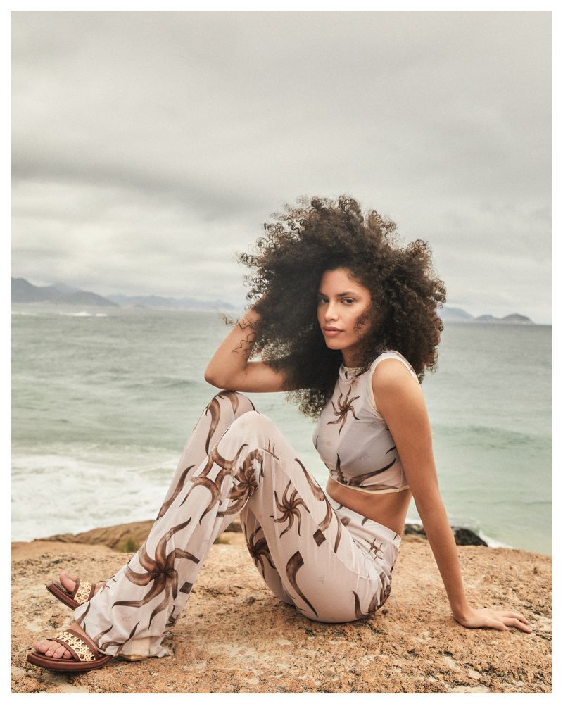 A woman with afro-style hair sits on a rocky outcrop by the ocean, wearing a patterned two-piece outfit and sandals in an outdoor setting.