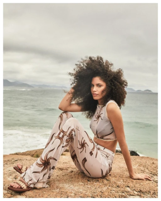 A woman with afro-style hair sits on a rocky outcrop by the ocean, wearing a patterned two-piece outfit and sandals in an outdoor setting.