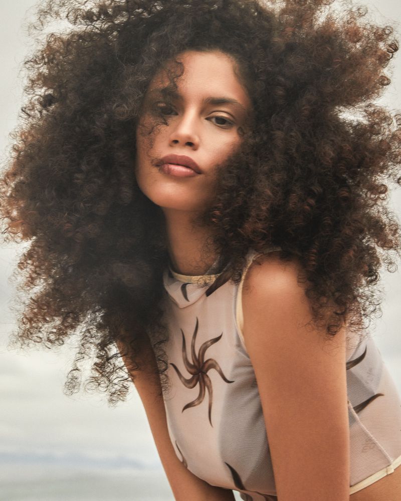 Woman with afro posing in sleeveless top with black sun pattern against bright sky, medium shot, fashion or beauty context.