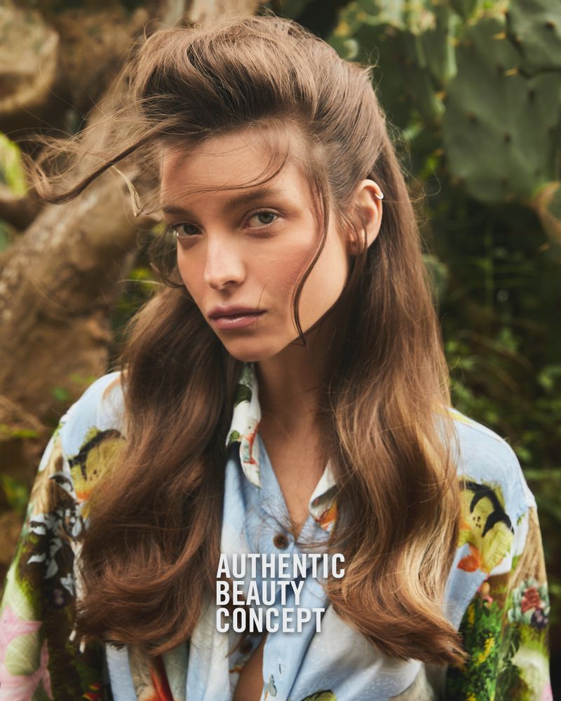 Brunette woman gazes at camera outdoors in diffused sunlight, wearing patterned shirt, with green leafy plants in soft-focus background.