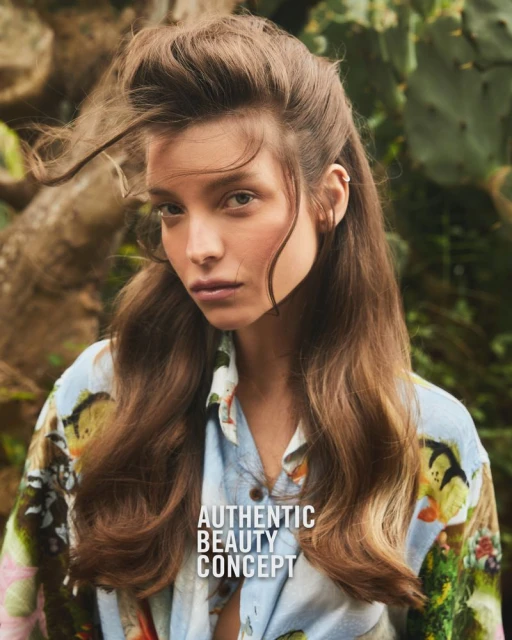 Brunette woman gazes at camera outdoors in diffused sunlight, wearing patterned shirt, with green leafy plants in soft-focus background.