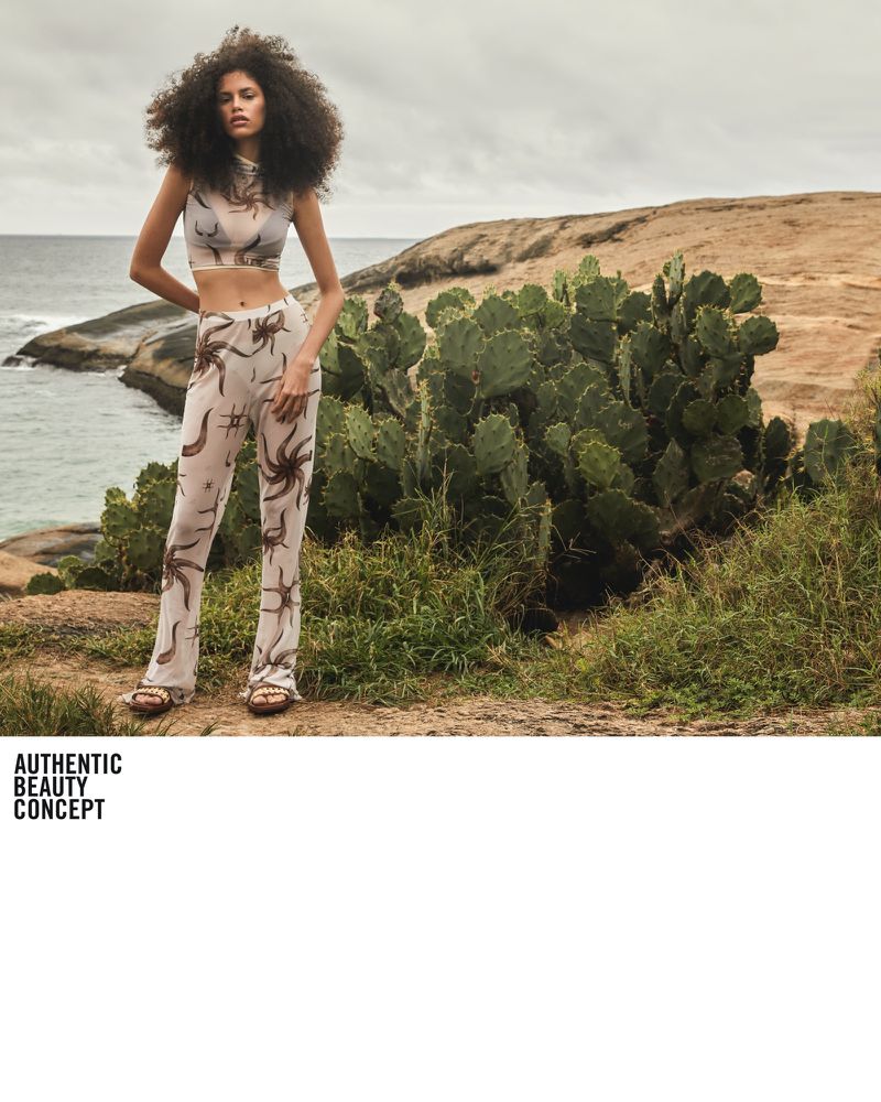 A young woman with afro hairstyle wearing a patterned two-piece outfit poses outdoors on a rocky hill overlooking the ocean.