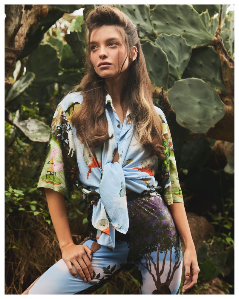 A brunette woman poses in a printed shirt and pants outdoors against lush foliage and cactus, lit with natural light.