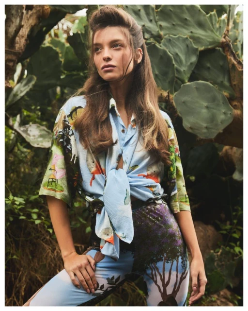 A brunette woman poses in a printed shirt and pants outdoors against lush foliage and cactus, lit with natural light.