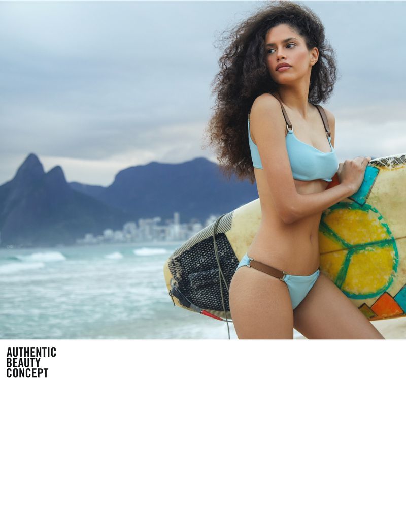 Adult woman in light blue bikini holding surfboard on beach with mountains in distance, wavy ocean and cloudy sky.
