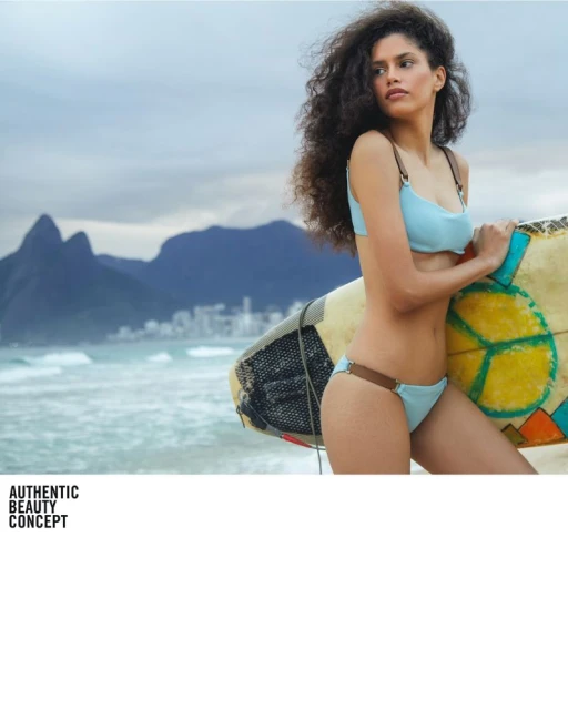 Adult woman in light blue bikini holding surfboard on beach with mountains in distance, wavy ocean and cloudy sky.