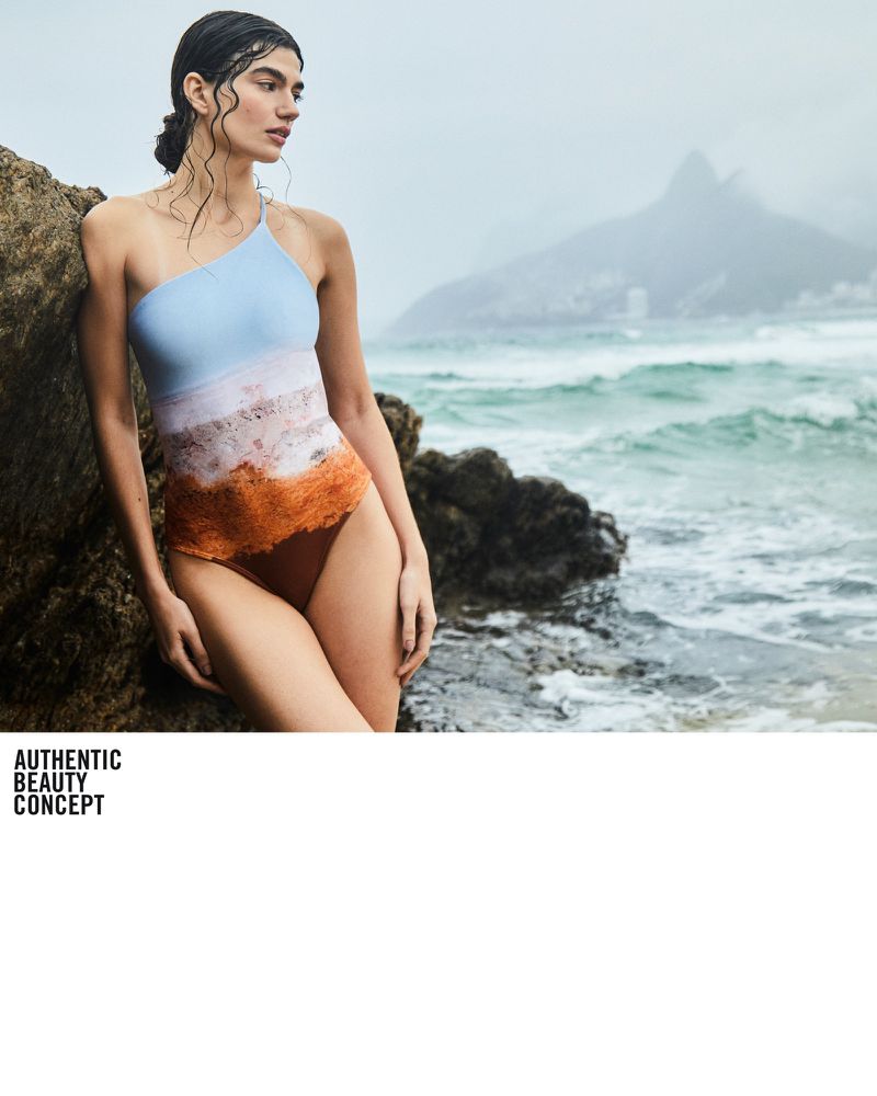 A woman poses in a one-shoulder swimsuit on a rocky shore with ocean waves and a mountain in the distance, showcasing a beach fashion aesthetic.