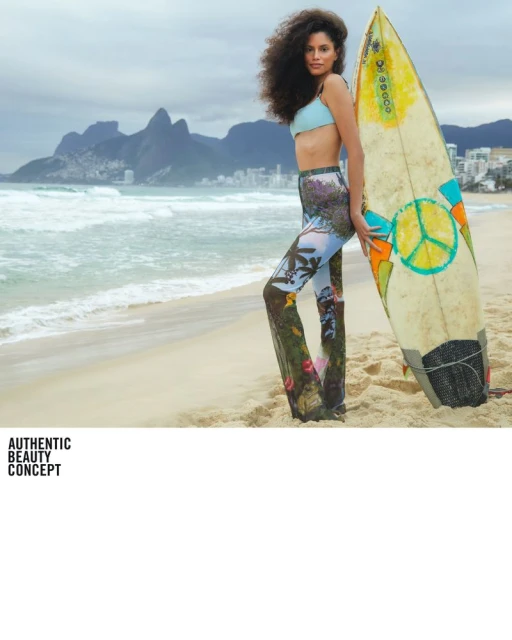 Woman posing with surfboard on a beach with mountains and cityscape visible in the background under an overcast sky.
