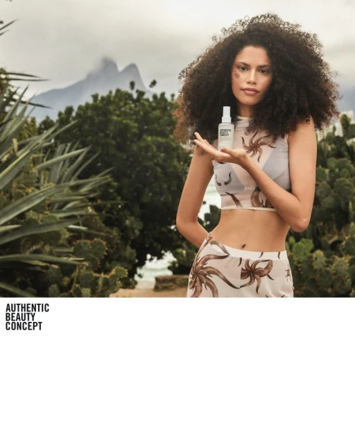 Woman presents beauty product against outdoor backdrop of tropical plants and mountains. Focus on the product and model's natural beauty.