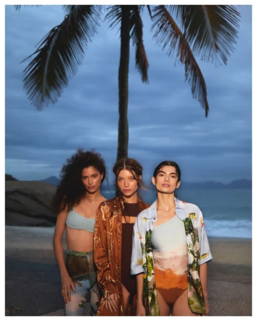 Three women in swimwear and beach cover-ups pose on a beach beneath a palm tree. The overcast sky adds a muted tone.