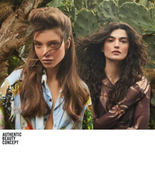 Two women with long brown hair pose outdoors amid lush greenery. One wears an open light blue shirt, the other a sheer brown top.