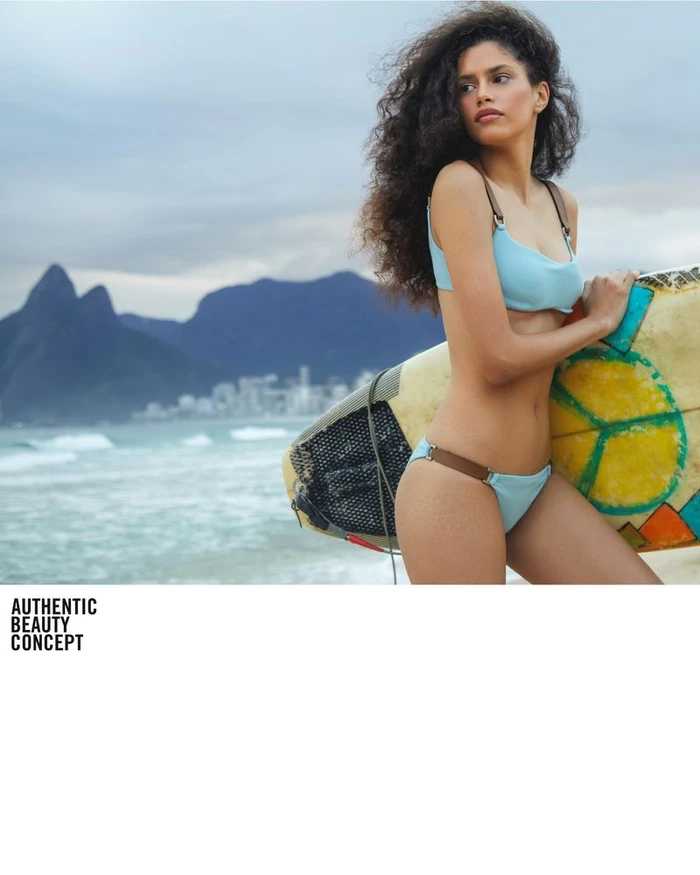Woman in a light blue bikini holding a surfboard on a sandy beach with mountains in the background