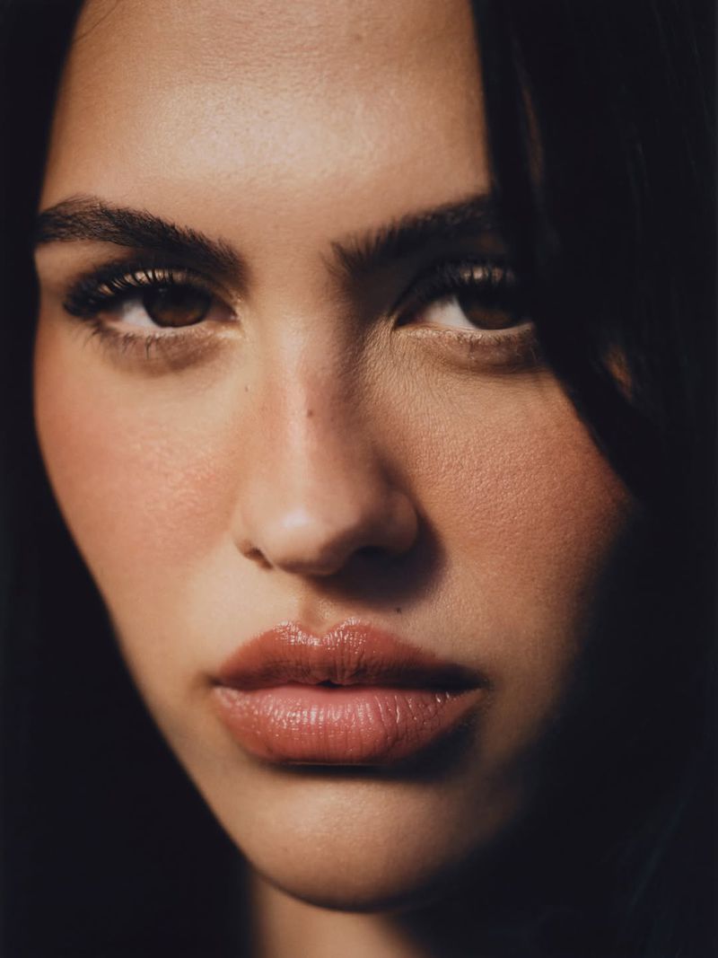 Close-up portrait of a woman with brown hair, brown eyes, and pink lipstick, set against a dark background with soft, diffused lighting.