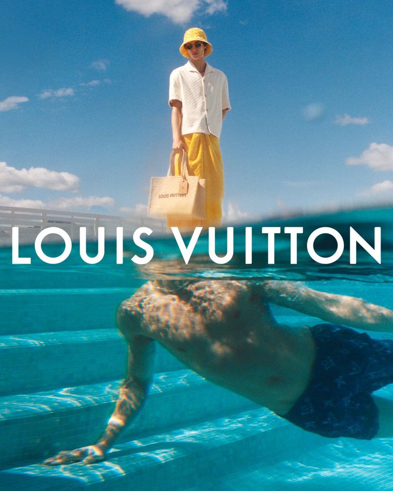 Fashion shoot with a man above water holding a tote bag and a swimmer below water, with a bright blue sky and turquoise water.