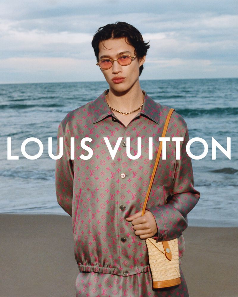 An adult male model poses on a beach with ocean waves in the background, wearing a patterned jacket and carrying a small woven purse.