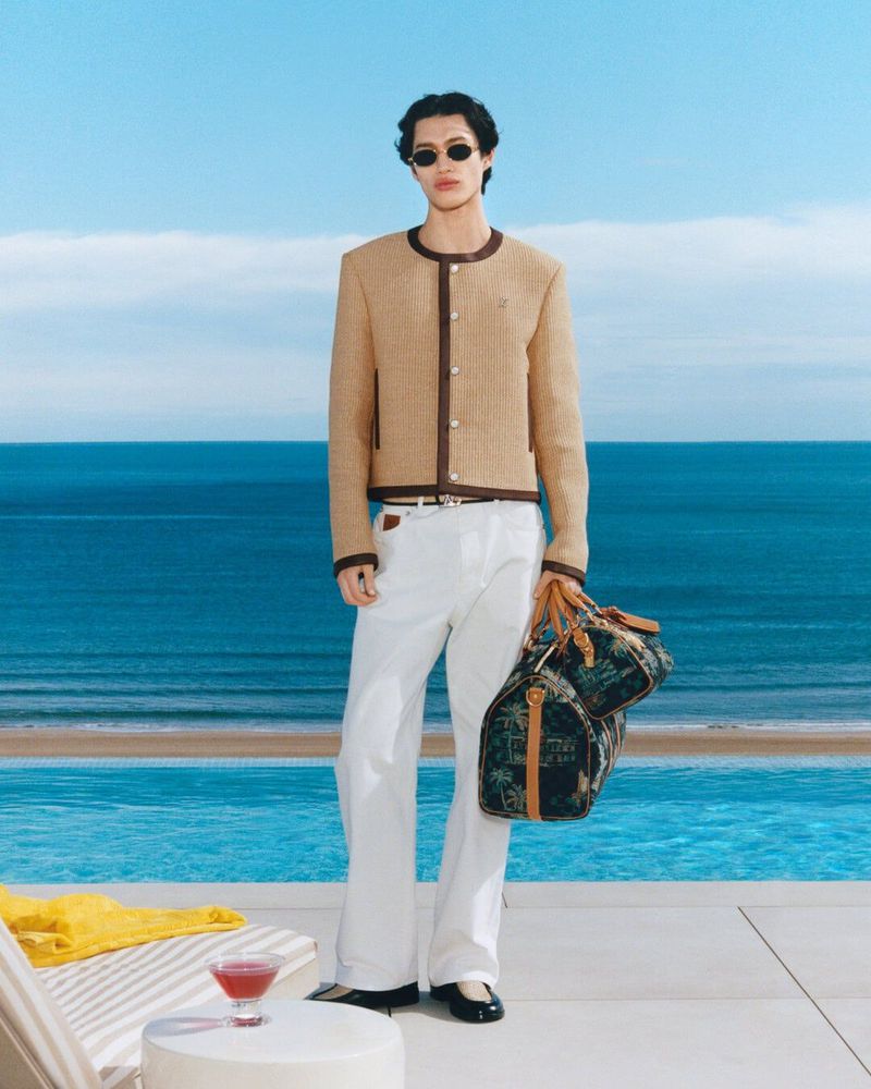 Adult male posing near an infinity pool, wearing a tweed jacket, sunglasses, and white slacks while holding a duffel bag.