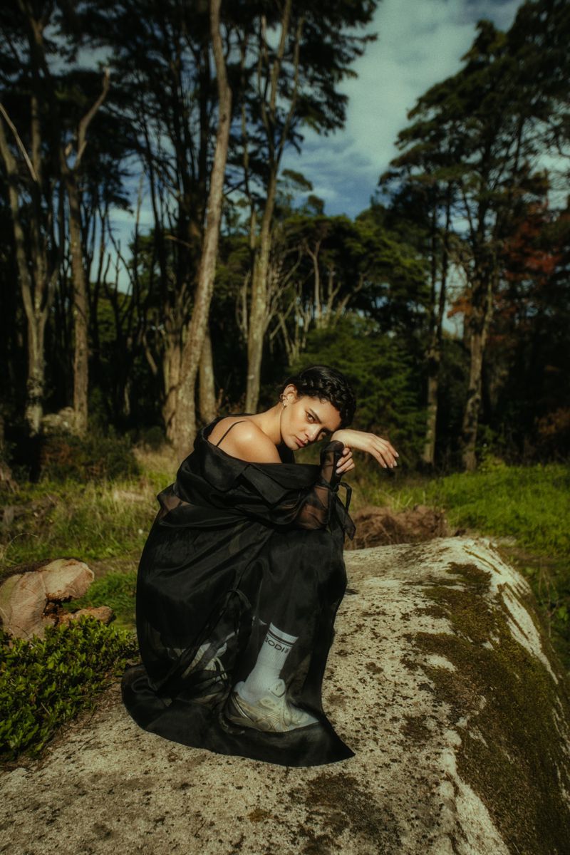A woman in a flowing black robe poses on a mossy rock in a forest, surrounded by tall trees, wearing sneakers and socks, with a somber expression.
