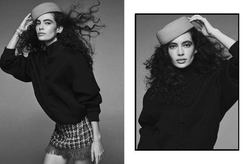 A black and white studio shot of a woman posing with curly hair, wearing a hat, black shirt, and tweed skirt with fringe.