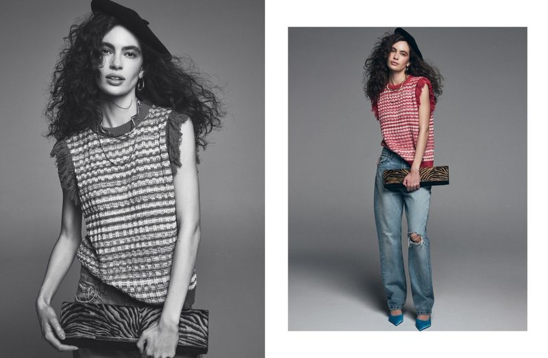 A woman wearing a knit top, wide-leg jeans, heels, and a hat poses in a photo studio with a grey backdrop, holding a clutch purse.