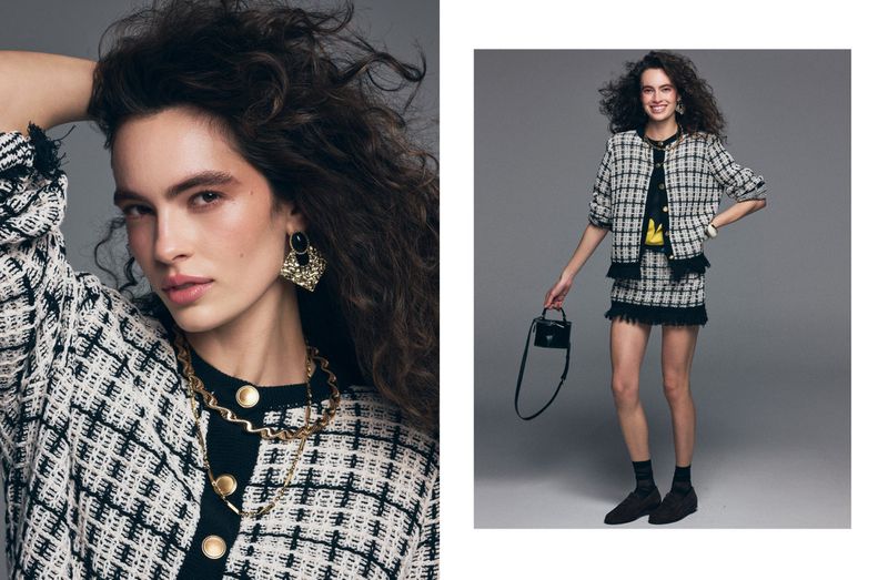 Fashion shot of a woman with curly brown hair posing in a black and white jacket and skirt, accessorized with gold jewelry.