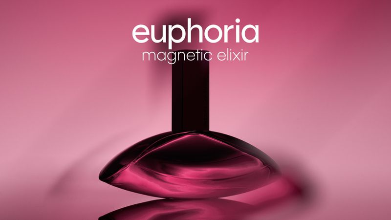Close-up studio shot of Calvin Klein's Euphoria Magnetic Elixir perfume bottle on a reflective surface against a pink gradient backdrop.