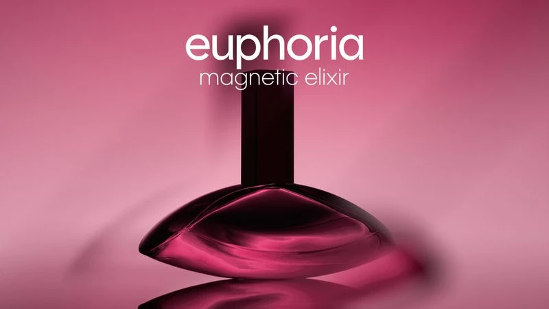 Close-up studio shot of Calvin Klein's Euphoria Magnetic Elixir perfume bottle on a reflective surface against a pink gradient backdrop.