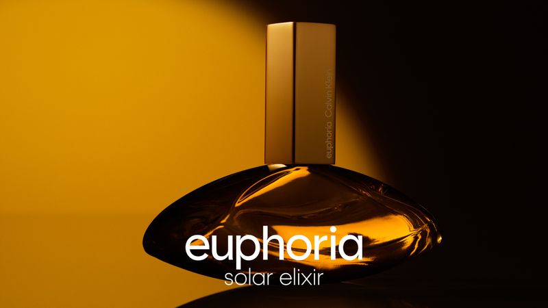 Gold Calvin Klein Euphoria Solar Elixir perfume bottle on a dark surface in a studio with a yellow background and soft lighting.