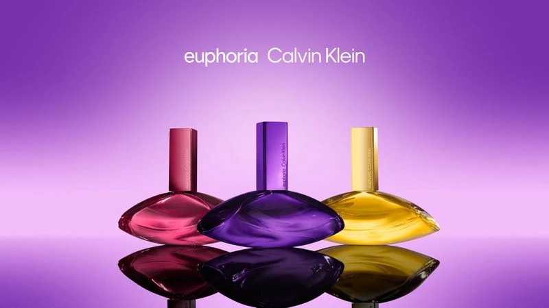 Three Calvin Klein Euphoria perfume bottles in pink, purple, and gold, lined up on a reflective surface against a gradient purple background.