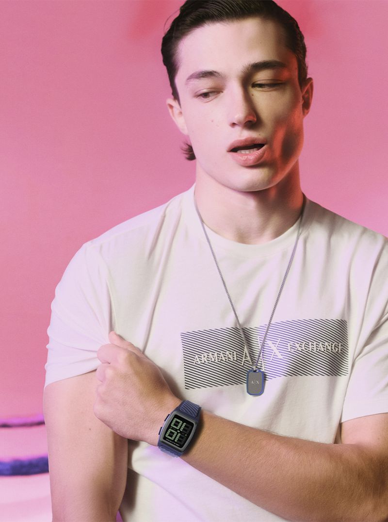 A young adult male in a white t-shirt, wearing a watch and necklace, poses against a vibrant pink background in a medium shot.