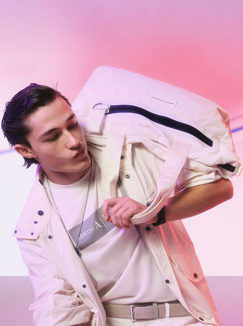 Stylish young man in off-white jacket and t-shirt, holding a matching bag, posing against a pink-lit backdrop. Fashion editorial shot.