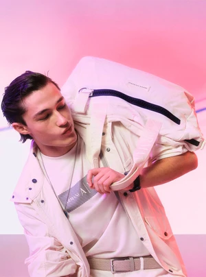 Male model carrying a large white duffel bag over his shoulder wearing a white jacket and t-shirt against a pink gradient background.