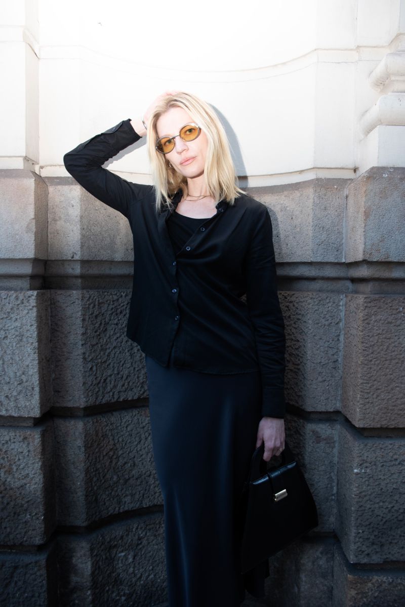 Blonde woman in sunglasses and black clothing poses against a stone wall, holding a black purse. Fashion editorial style portrait.