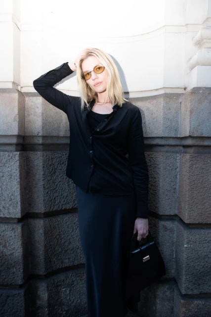 Blonde woman in sunglasses and black clothing poses against a stone wall, holding a black purse. Fashion editorial style portrait.