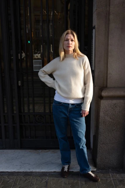 Adult woman in an off-white knit sweater and blue jeans stands in front of a dark wrought iron gate of a stone building.