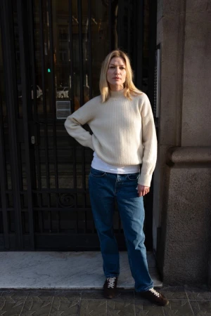 Adult woman in an off-white knit sweater and blue jeans stands in front of a dark wrought iron gate of a stone building.