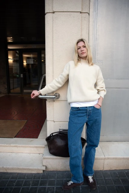 Blonde woman in a cream sweater and jeans, posing casually against a stone wall with a brown bag and steps visible.
