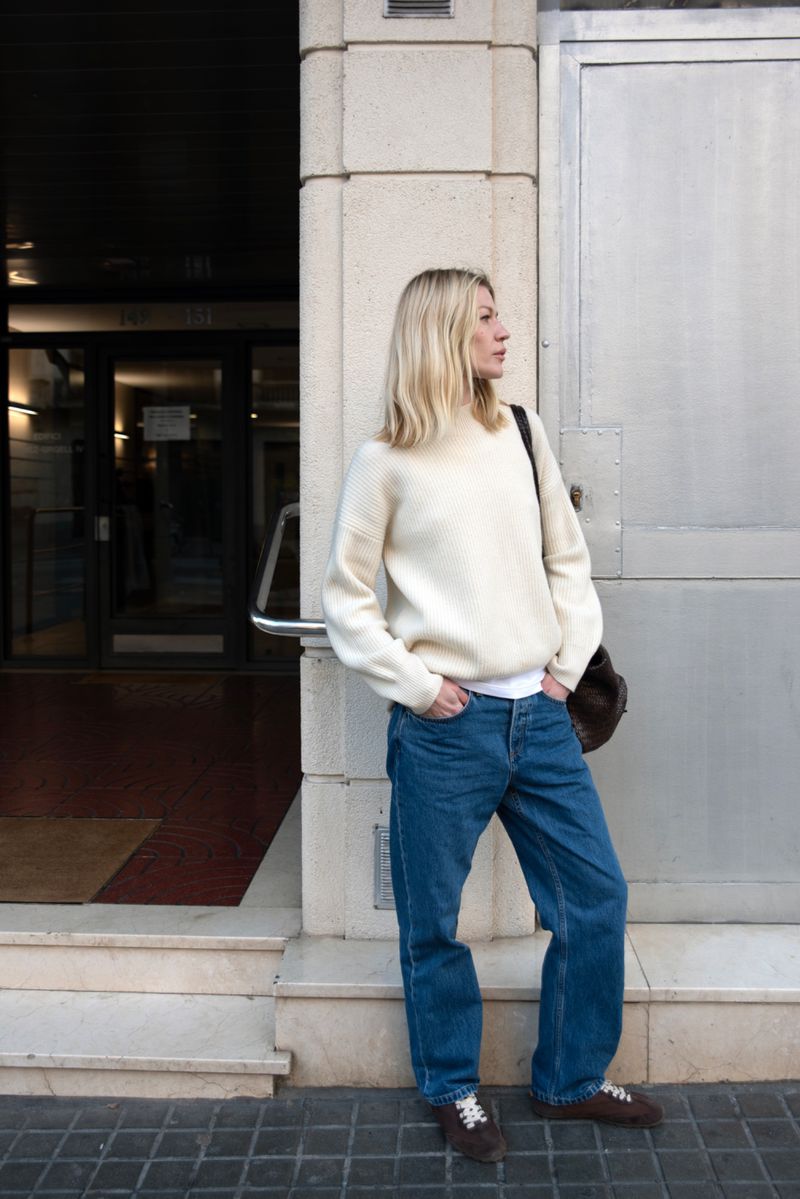 Blonde woman in cream sweater and blue jeans standing outside a building exterior for Asket advs editorial photography.