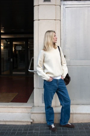 Blonde woman in cream sweater and blue jeans standing outside a building exterior for Asket advs editorial photography.