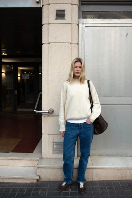 Blonde woman stands outside a building in blue jeans and a cream sweater with a brown tote bag, looking at the camera.