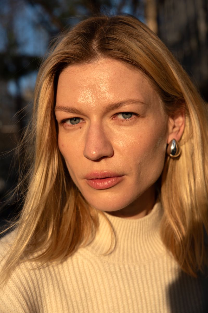 Close-up portrait of a blonde woman with light skin and blue eyes, wearing a cream turtleneck sweater and silver earrings outdoors.