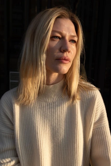 Portrait of a blonde woman with fair skin wearing a cream-colored sweater. Soft light illuminates her face against a dark background.