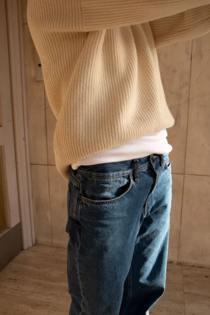 A person in blue jeans and an off-white ribbed sweater stands indoors, illuminated by warm light highlighting the textures of the fabric.