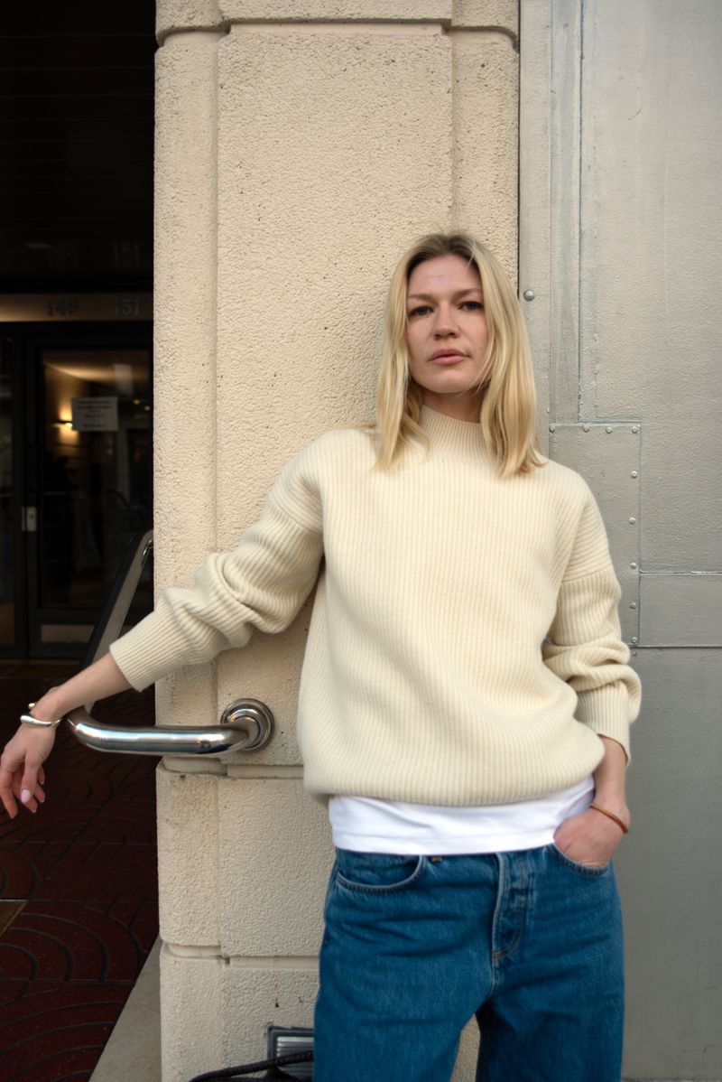 Blond woman poses outdoors wearing a cream-colored sweater, blue jeans, and a white t-shirt; leaning against a cream-colored wall.