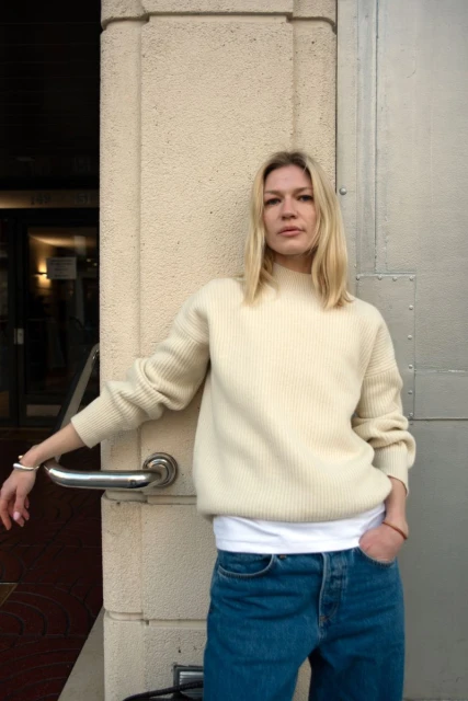Blond woman poses outdoors wearing a cream-colored sweater, blue jeans, and a white t-shirt; leaning against a cream-colored wall.