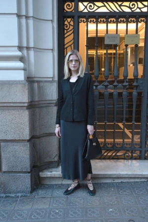 Woman wearing a black button-down shirt and long black skirt holding a black handbag outside a building entrance