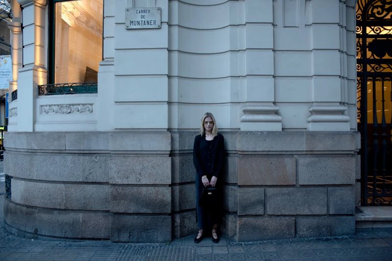A woman stands on a city street in front of a white stone building, wearing a black jacket and holding a black handbag in an urban setting.