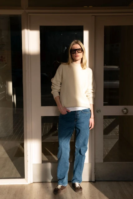 Adult female wearing a cream sweater, jeans, and sunglasses stands outside a door in the sunlight, posing for a fashion shot.