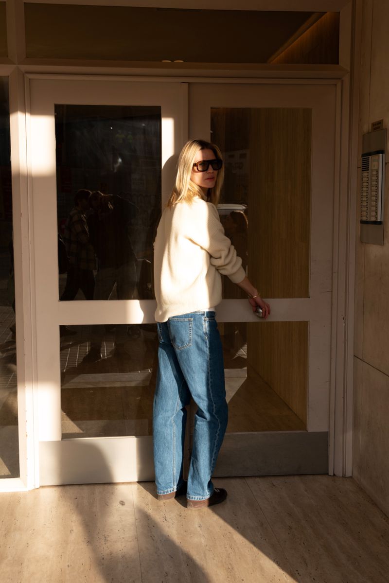 Adult woman with blonde hair, wearing a casual sweater and jeans, exits a building through glass double doors with sunlight hitting the frame.